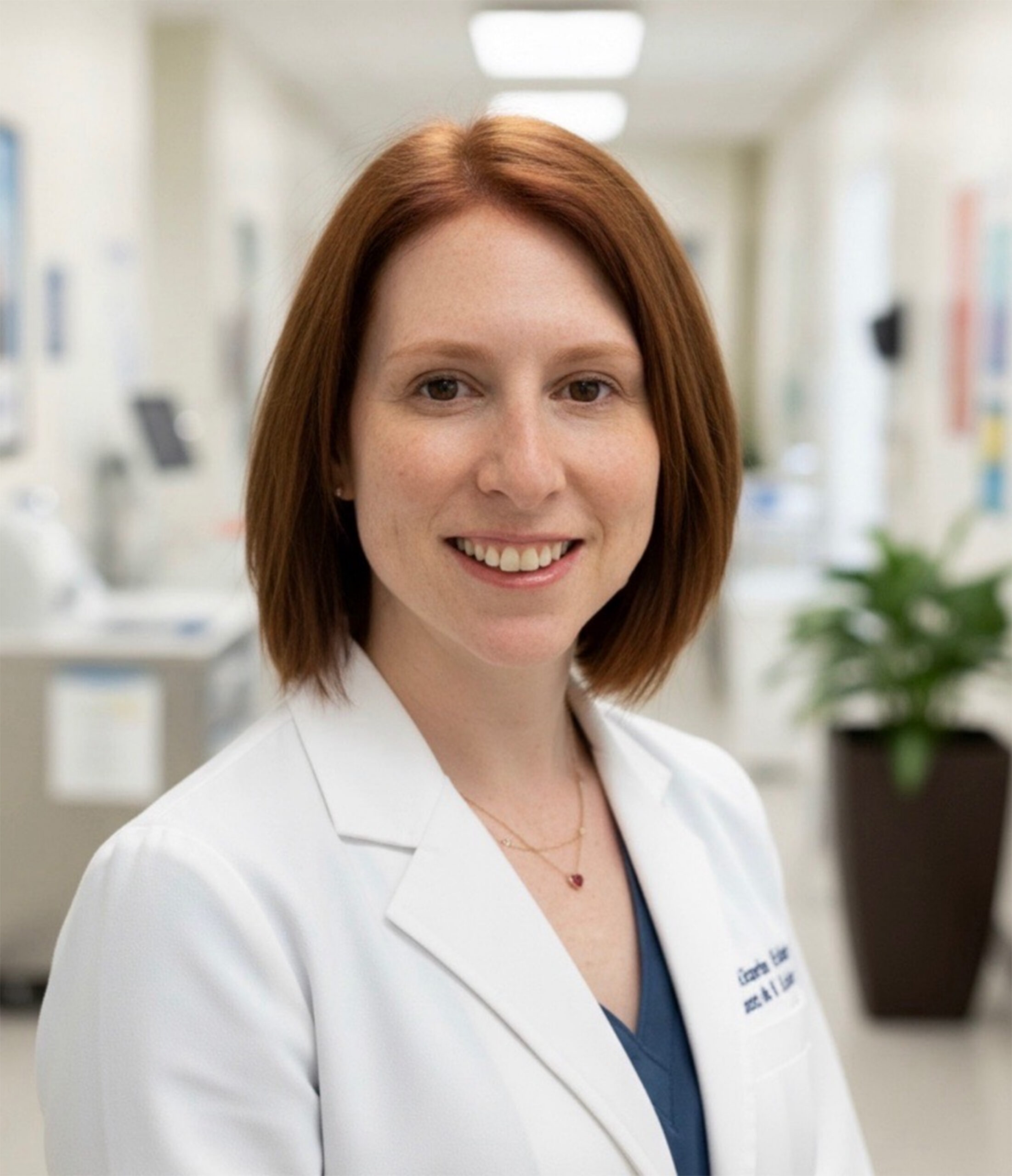 Smiling female healthcare professional in a white lab coat in a bright clinic corridor, head and shoulders visible.